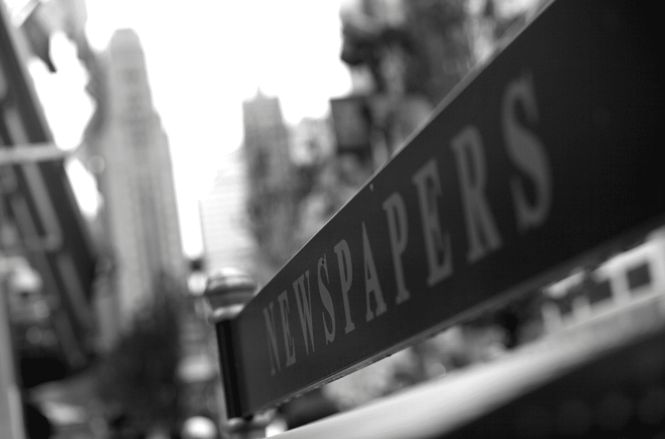 A black and white photo with a sign that says "newspapers" on it. In the background, there are blurred images what appear to be city skyscrapers. 